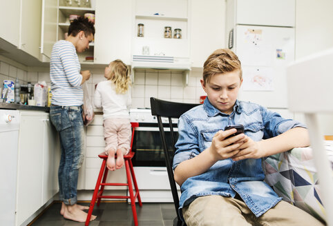 Boy using mobile phone in kitchen at home with mother and sister in background - stock photo